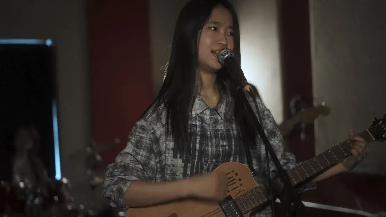 The band's vocalist sings into a microphone and plays an acoustic-electric guitar in a dim rehearsal room, with the drummer blurred behind her