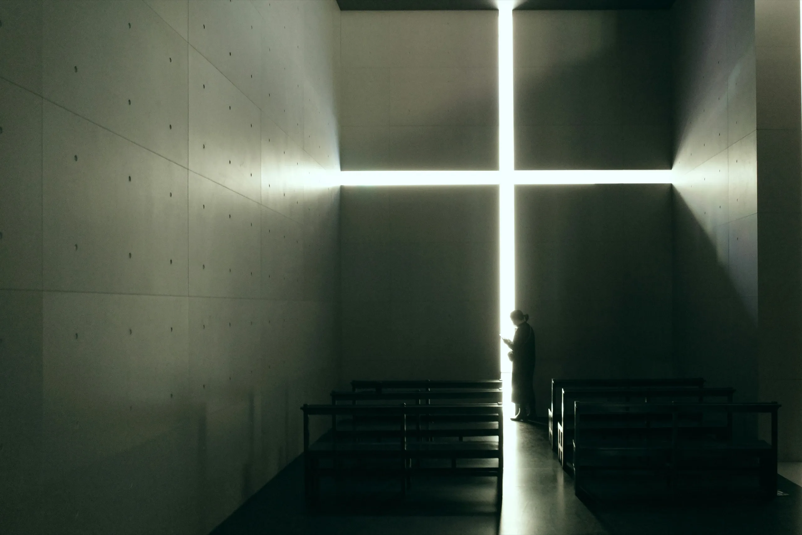 A cross of light cut into a concrete chapel wall, with one person standing beside the pews