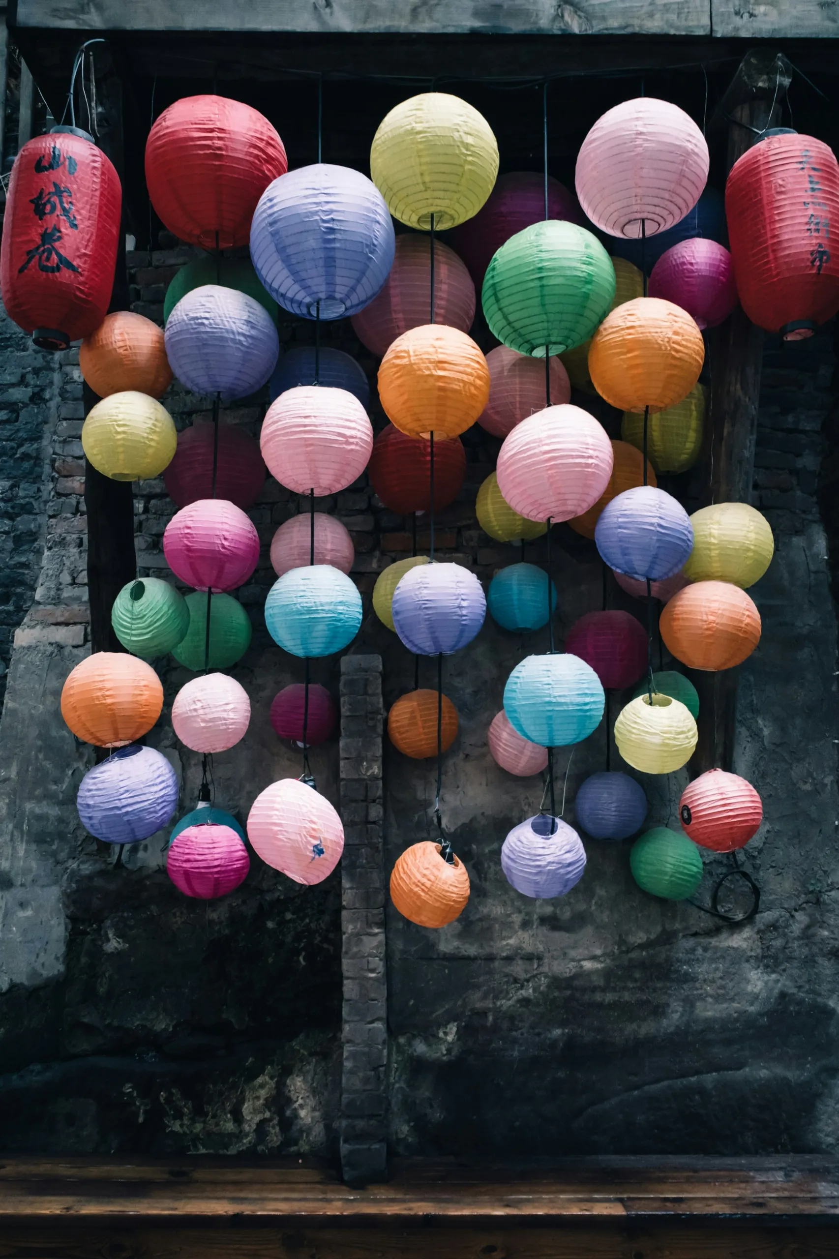 Paper lanterns in many colours hanging against a dark brick wall