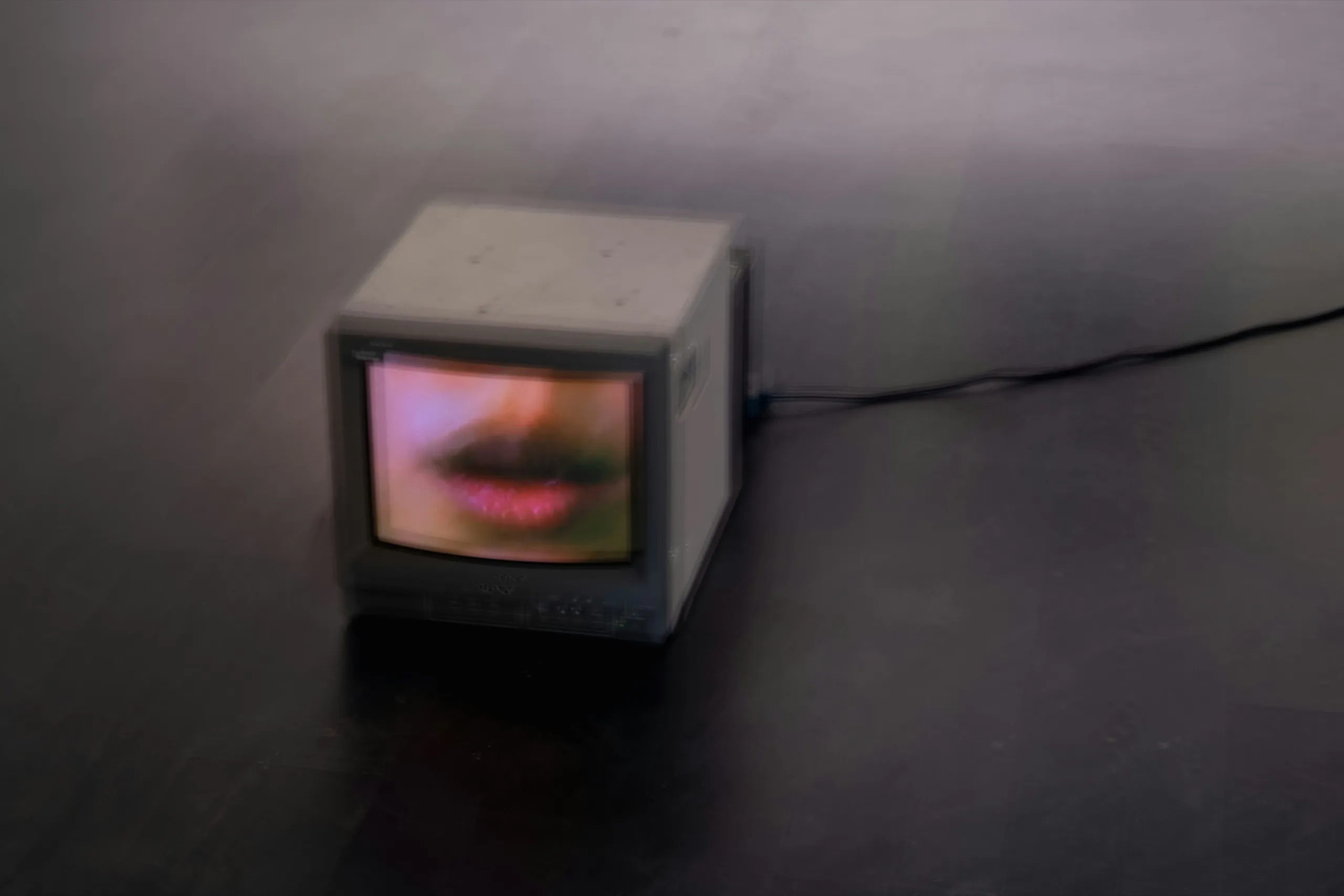 A small grey television on a dark floor, its screen filled with a close-up of lips